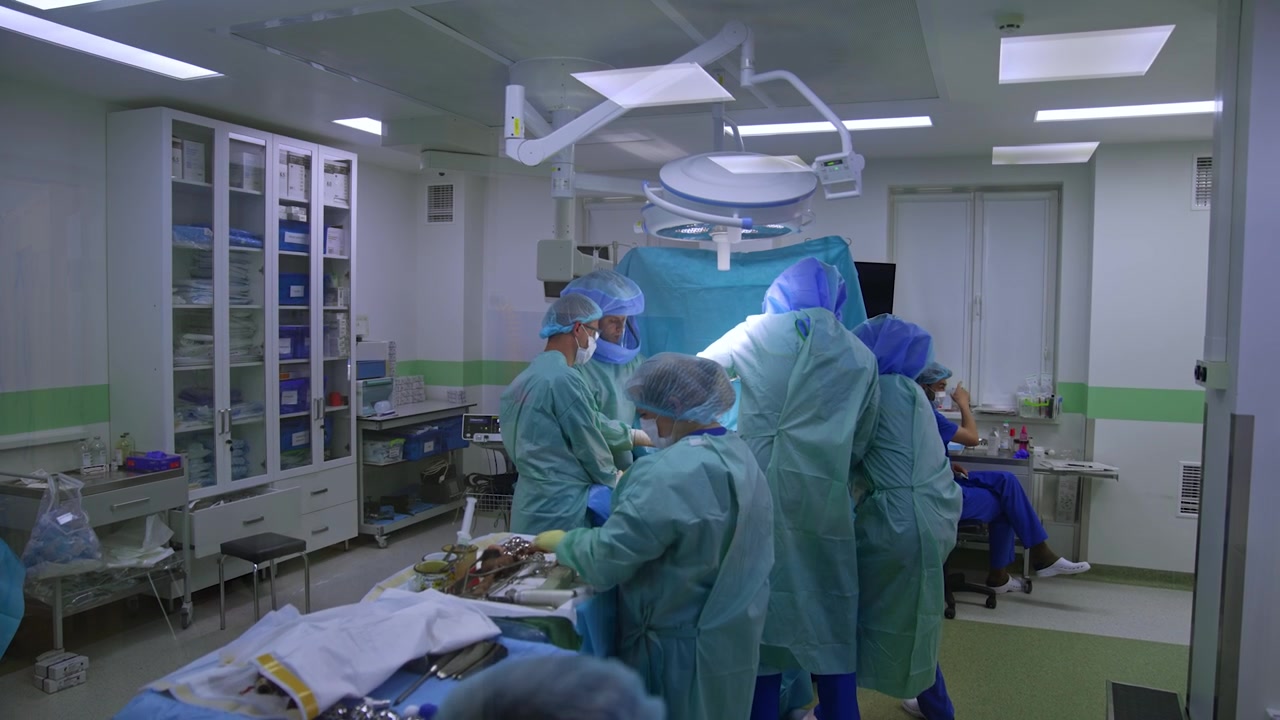 Nærmer sig den lysegrønne skydedør med et vindue i. Et stort lægeteam udfører en operation inde i rummet.
