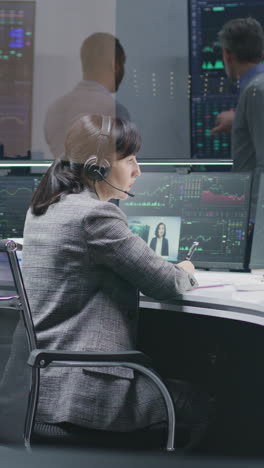 Female-financial-analyst-and-investment-manager-discuss-real-time-stocks-data-on-computer.-Colleagues-work-in-bank-office.-Big-digital-screens-showing-exchange-market-charts.-Vertical-shot