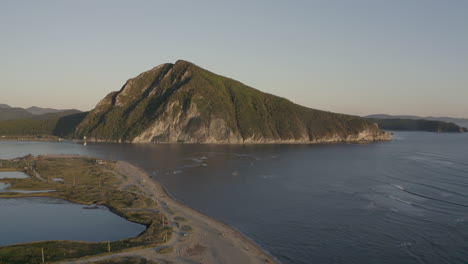 a landscape view of a pyramid-shaped mountain located at a river estuary flowing into the sea, with green vegetation on its sides, mountain ridge in the background, on the sunset