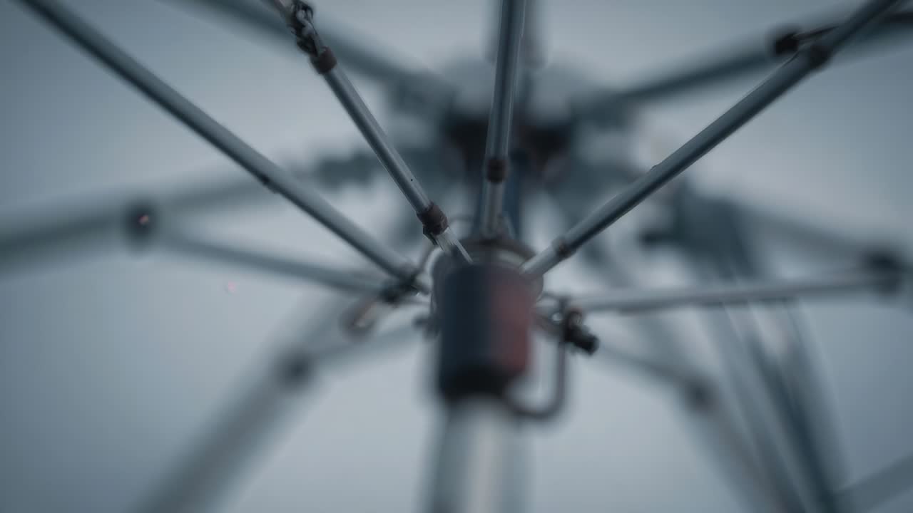 Opening umbrella hub extending metal ribs under pale overcast sky, with droplets for shelter