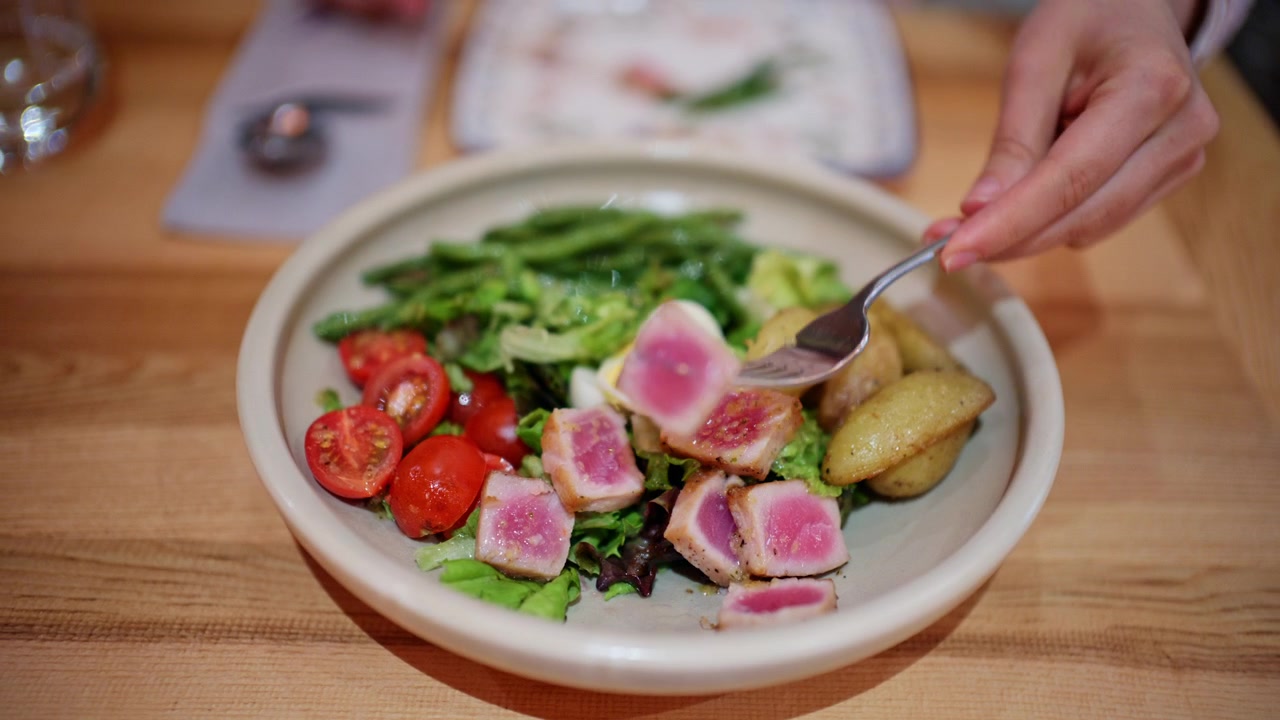 A bowl of salad placed on a wooden table, containing leafy greens, cherry tomatoes, boiled egg slices, and pieces of seared tuna