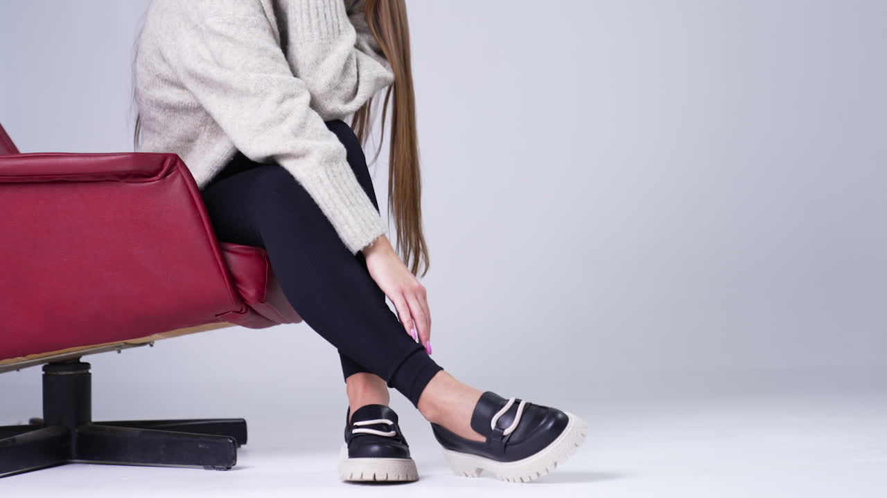 Female sitting in a red chair stretching hand to her shoes. Modern stylish footwear demonstration in the studio. White backdrop.