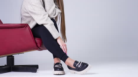 female sitting in a red chair stretching hand to her shoes. modern stylish footwear demonstration in the studio. white backdrop.
