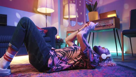 young stylish caucasian man with long hair in headphones lying on the floor and using smartphone in a retro party at home