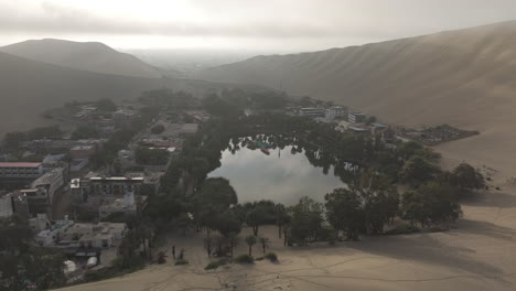 drone shot flying over the city of huacachina in peru behind the dunes on a cloudy day during the mist in the morning with the sun coming through log