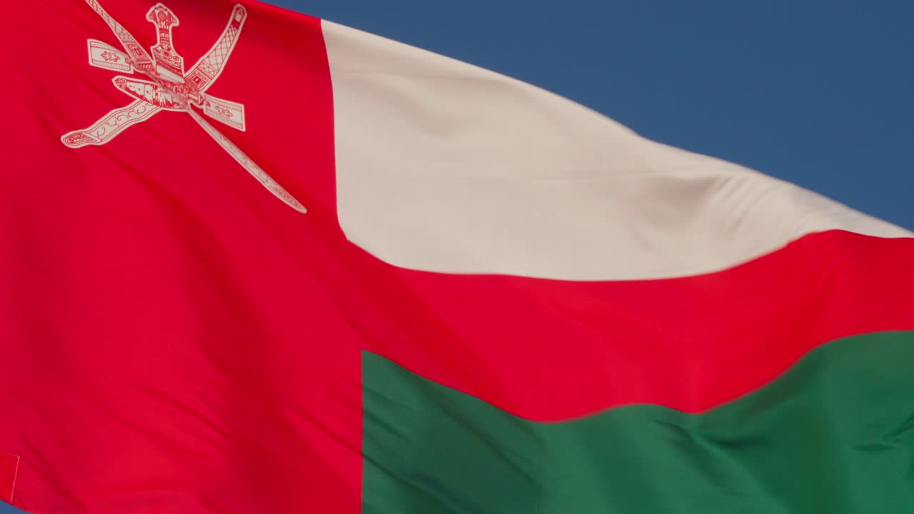 Flag of Oman featuring national emblem and horizontal red, white, and green stripes waving close-up. Location: Muscat, Oman