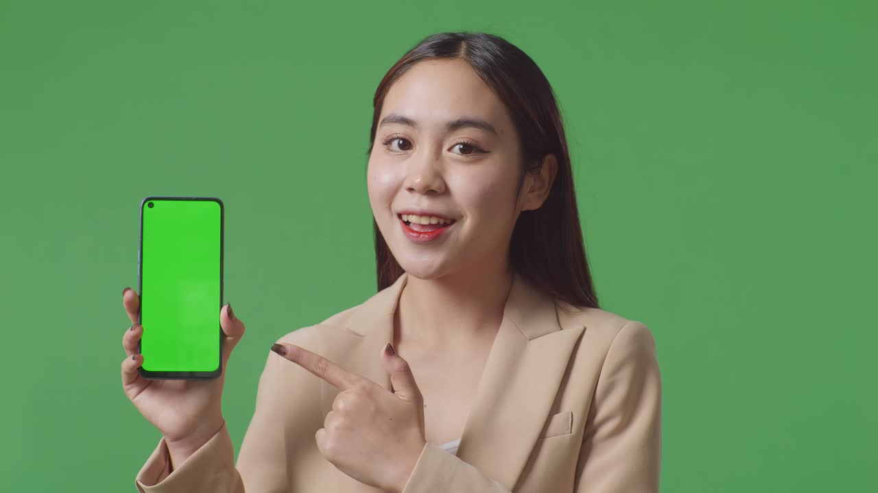 초록색 화면 앞에 서 있는 동안 녹색 화면 휴대 전화를 보여주고 가리키는 미소를 짓는 아시아 사업가 여성의 클로즈업