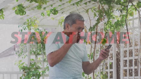 Quédese-En-Casa-Texto-Contra-Un-Hombre-Afroamericano-Tomando-Café-Y-Usando-Un-Teléfono-Inteligente-En-El-Jardín