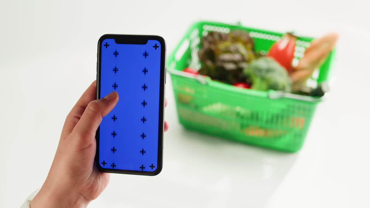 Person holding a smartphone with a blue screen in front of a shopping basket filled with groceries.