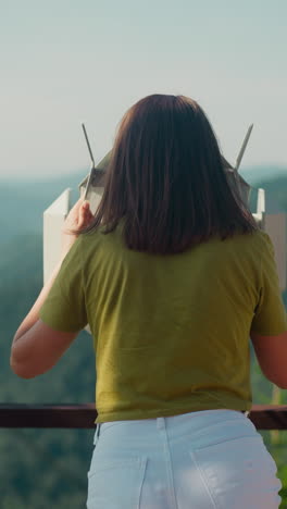 woman with loose brunette hair stands at top of mountain looking through binoculars. lady uses huge binoculars for better contemplation of mountainscape