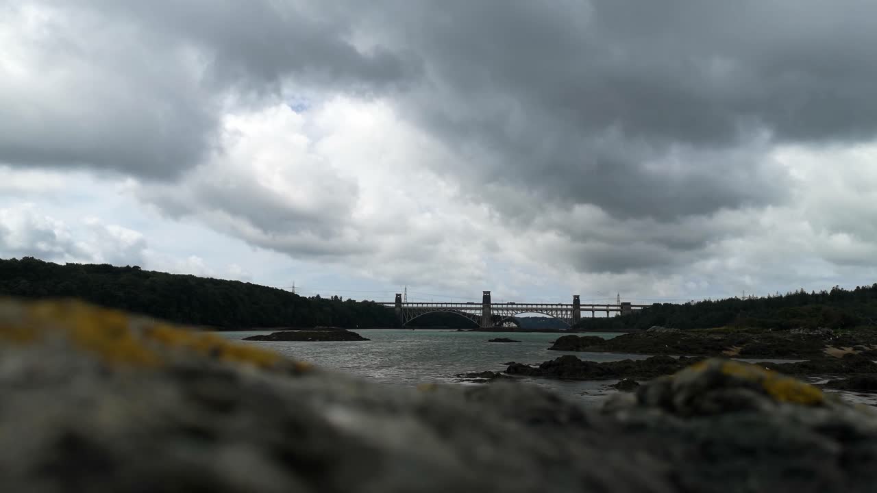 глядя на нефокусированную скалу на переднем плане к облачному проливу менай британский мост к англеси