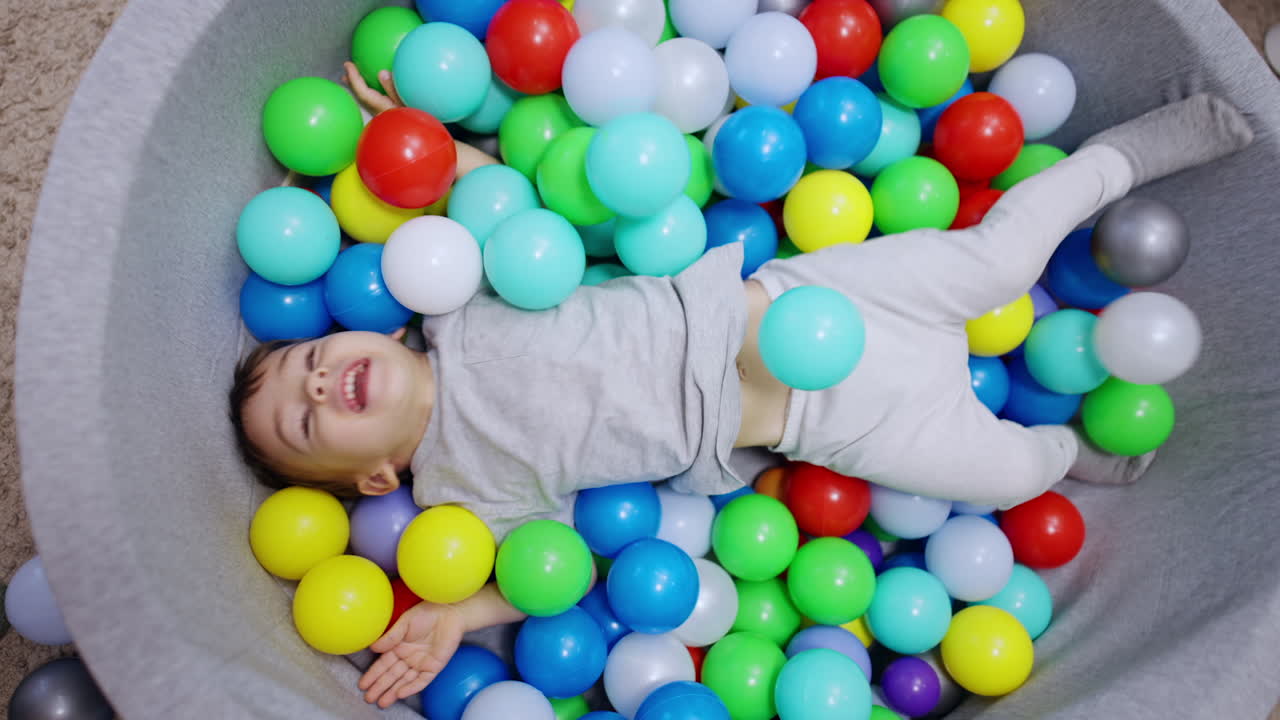 Playful Caucasian toddler lies in the dry basin. Smiling kid waves his hands and feet among the balls. Top view.