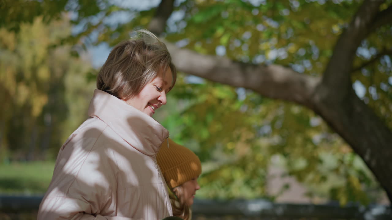 Young mom strolls side by side with son through vibrant autumn park, both glancing toward distant scene as mom holds dog leash loosely, golden foliage overhead and sunlit pathway