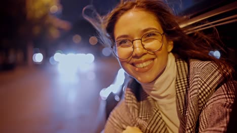 A-happy-and-cheerful-brunette-girl-in-round-glasses-smiles-broadly-in-a-gray-coat-and-looks-out-of-the-rear-window-of-a-modern-car-in-the-evening.-Happy-brunette-girl-looks-out-of-the-car-window-during-an-evening-drive-around-the-city