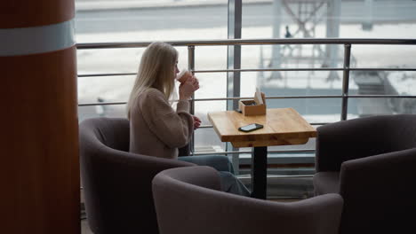 young lady holding coffee cup, sipping from it in a mall cafe with blurry city view through window, phone placed on table with serene atmosphere