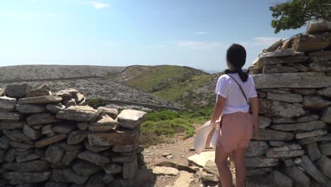 Back-view-of-girl-in-summer-hiking-outfit,-standing-in-front-of-stone-fence,-looking-into-distance