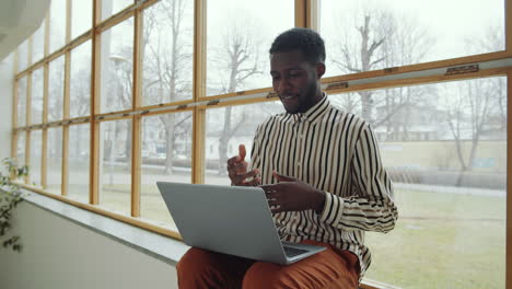 black man sitting on window sill and video calling on laptop