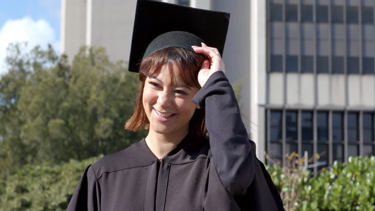 Donna in toga di laurea nera che tiene in mano un diploma arrotolato, sorridendo, sistemandosi il tocco di laurea nel campus