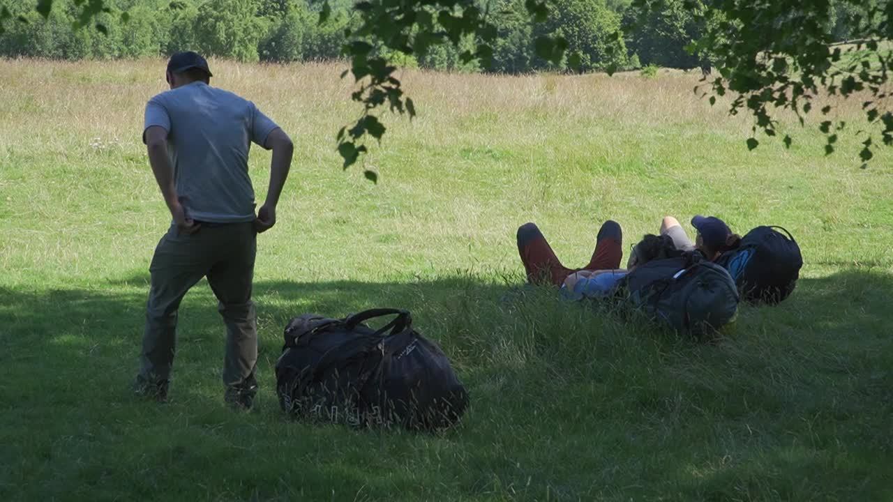 A man joins two others sitting in the shade, taking a moment to relax in a green, natural area.