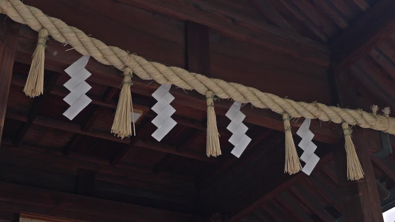 A traditional Shimenawa rope adorned with white Shide paper strips hangs beneath the eaves of a wooden structure at Kuramae Shrine.