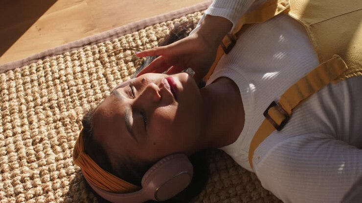 La lumière du soleil qui entre déclenche chez une femme asiatique l’envie d’ajuster ses écouteurs et de s’installer pour écouter de l’audio sur un tapis de jute