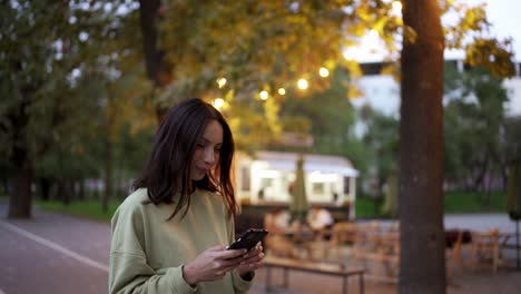 Brunette-girl-in-a-green-sweater-is-chatting-on-the-phone-against-the-backdrop-of-and-evening-park.-Cafe-in-the-park,-spending