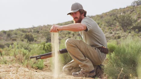 kneeling caucasian male survivalist holding hunting rifle, examining sandy terrain of wilderness