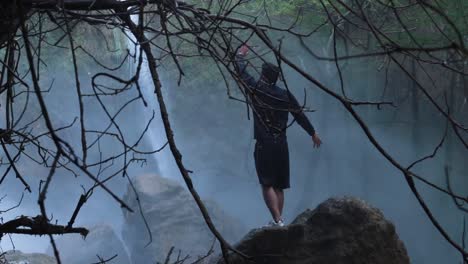 hombre joven, de 20 años, de pie sobre una roca frente al rocío de la cascada durante el día nublado, plano general