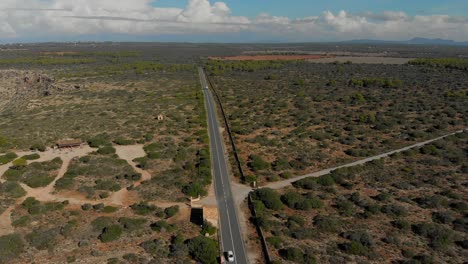 Drone-footage-flying-over-a-road-with,-cars-driving-on-it,-in-a-flat-landscape-covered-with-sand,-stones-and-bushes