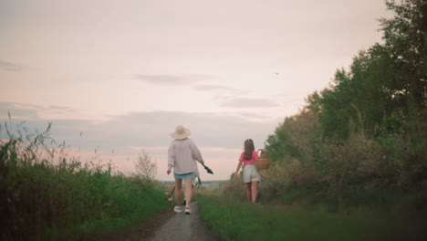 vista trasera de la familia caminando a casa a lo largo de un camino de tierra al atardecer, con un pájaro volando por encima, dos mujeres con sombreros caminando con un perro en una correa, mientras una niña joven lleva una canasta y un niño juega