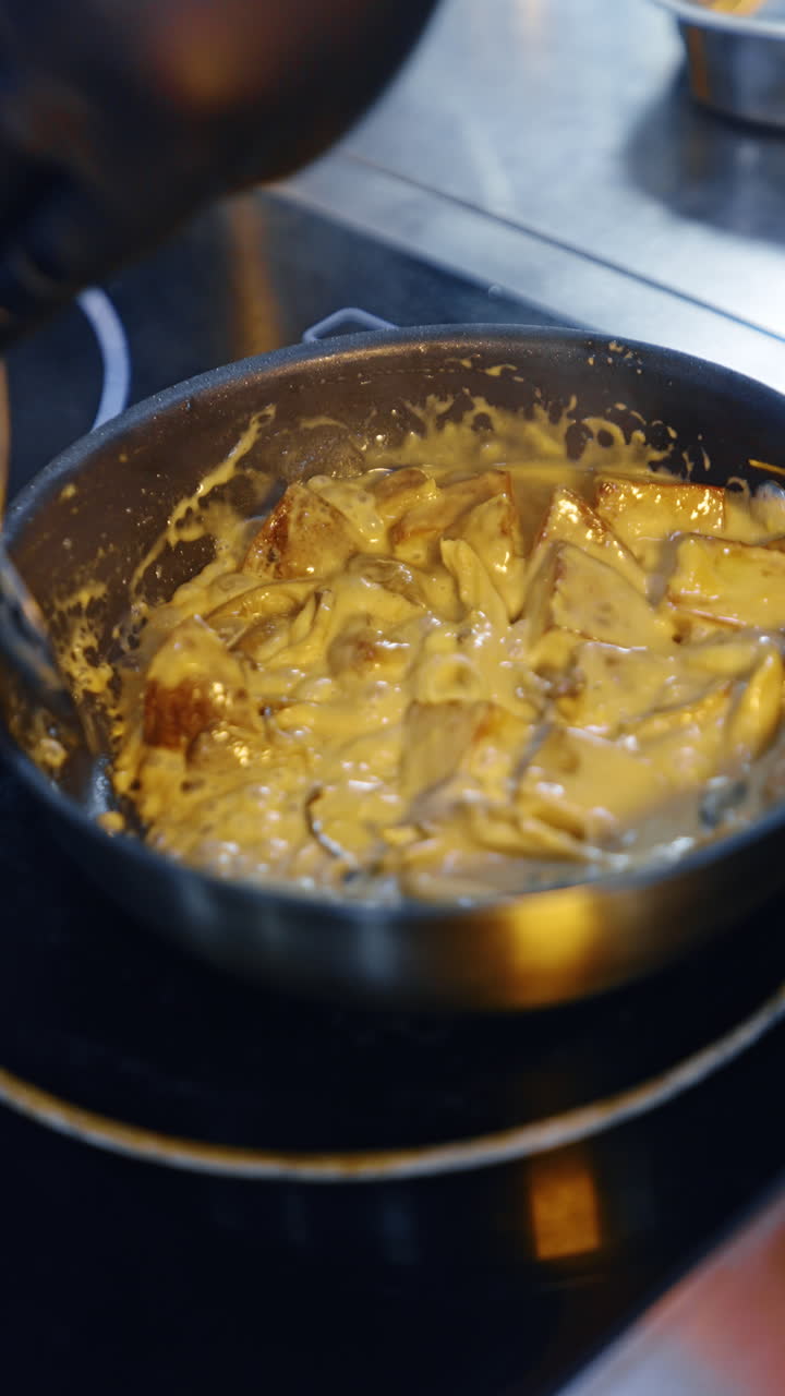 Preparing the potatoes in the cheese sauce. Cook stirs the dish in frying pan. Close up. Blurred backdrop. Vertical video.