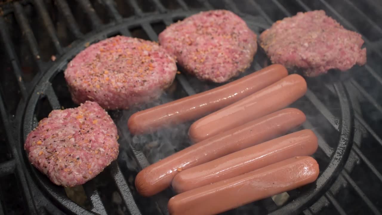 el primer plano de la parrilla al aire libre mientras las hamburguesas de carne de vacuno sazonadas crudas y los perritos calientes se cocinan sobre llamas con humo.