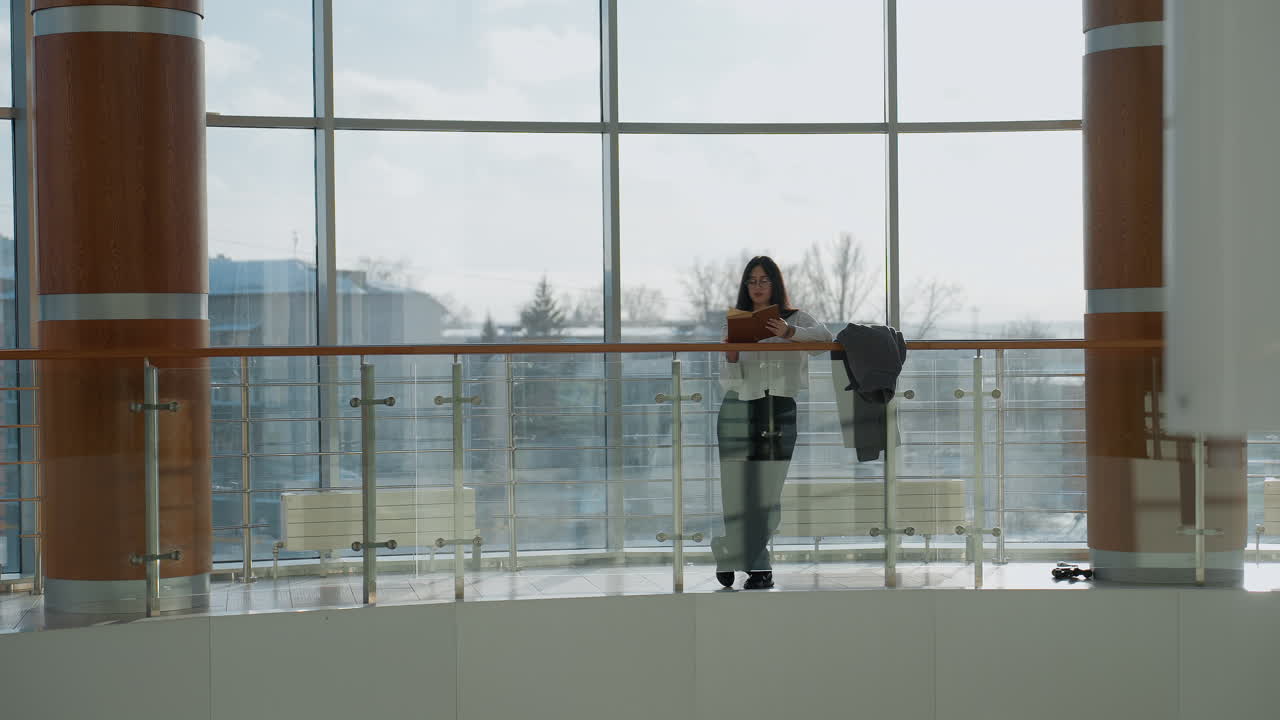 Confident lady stands relaxed, leaning on railing of indoor balcony with jacket hanging nearby, modern urban landscape and city architecture visible through large glass windows behind her