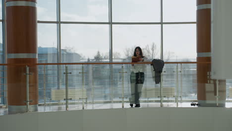 confident lady stands relaxed, leaning on railing of indoor balcony with jacket hanging nearby, modern urban landscape and city architecture visible through large glass windows behind her