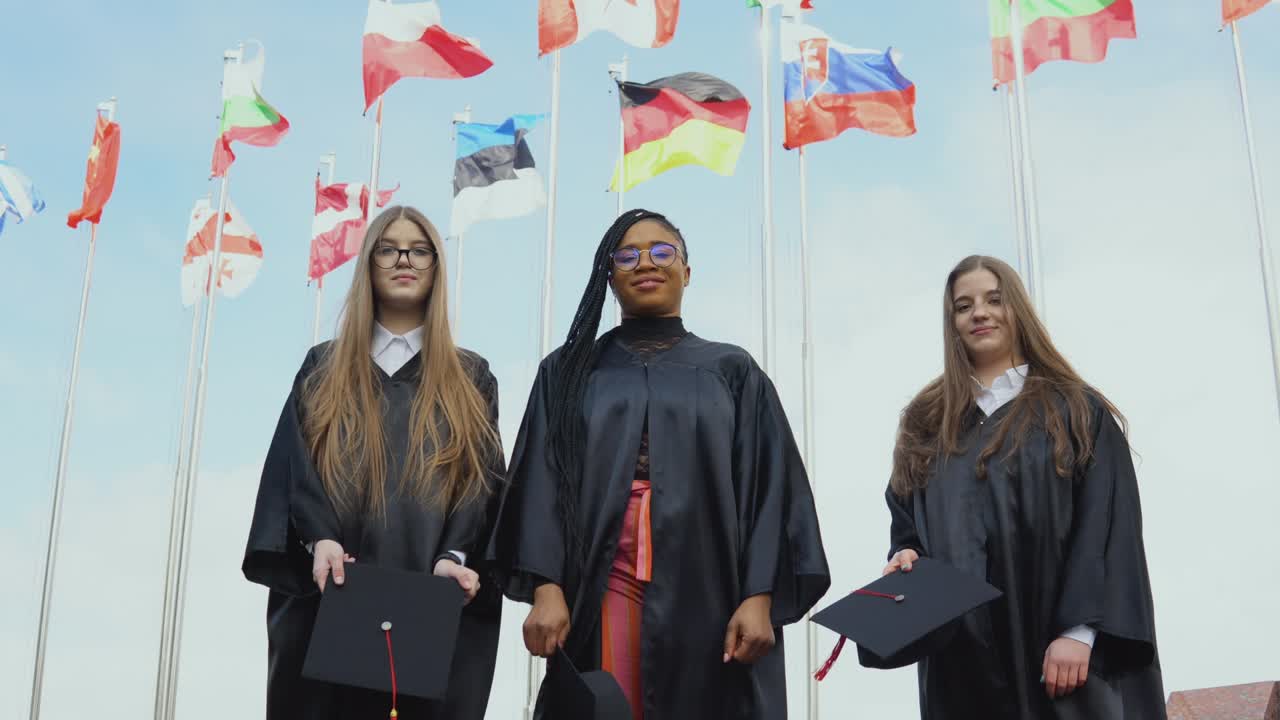 Three young women of different nationalities joyfully throw hats up against many flags of different countries fluttering in the wind on the background. Outdoor view from bottom