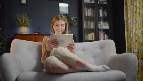 mujer joven sonriente con auriculares usando tableta mientras se sienta en el sofá en casa