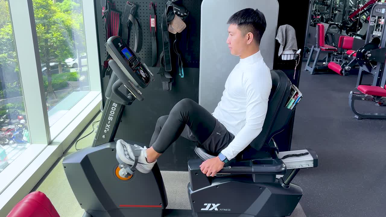 Young man exercises on recumbent bike in bright gym with large windows and natural light