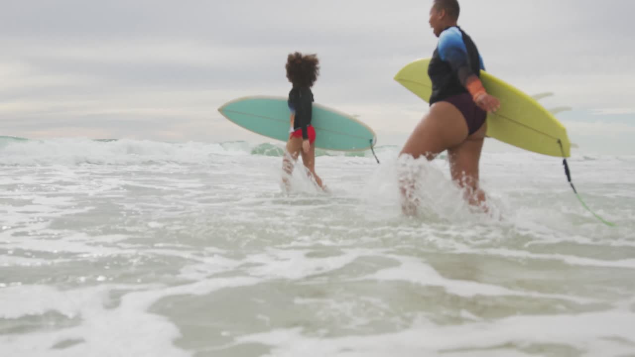 felices amigas afroamericanas corriendo hacia el mar sosteniendo tablas de surf