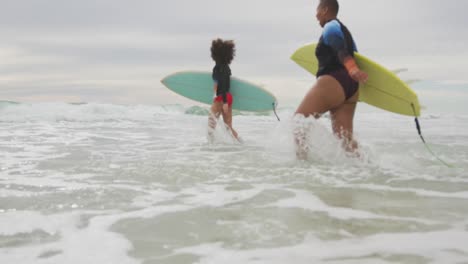 happy african american female friends running into the sea holding surfboards