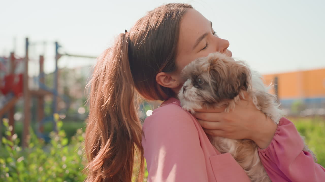 Young Girl In Pink Embracing Puppy Lovingly, Cheerful Girl With Ponytail Snuggling Puppy Near Park Fence, Woman With Bright Expression Hugging Her Puppy Close To Her Chest In Urban Park Setting