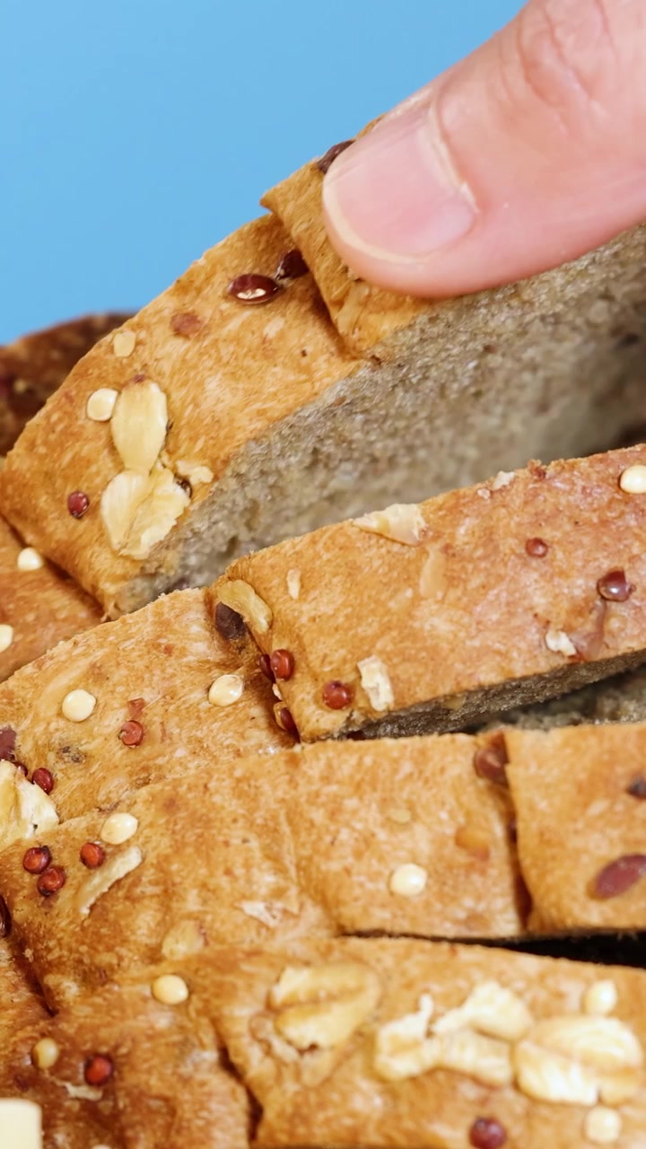A hand picks a slice of whole wheat bread from a stack, set against a bright blue backdrop