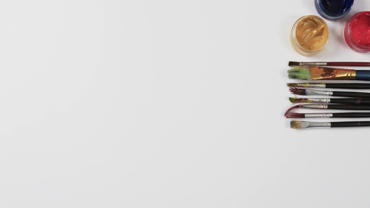 vista aérea de pinturas y pinceles dispuestos sobre un fondo blanco con espacio de copia, en cámara lenta
