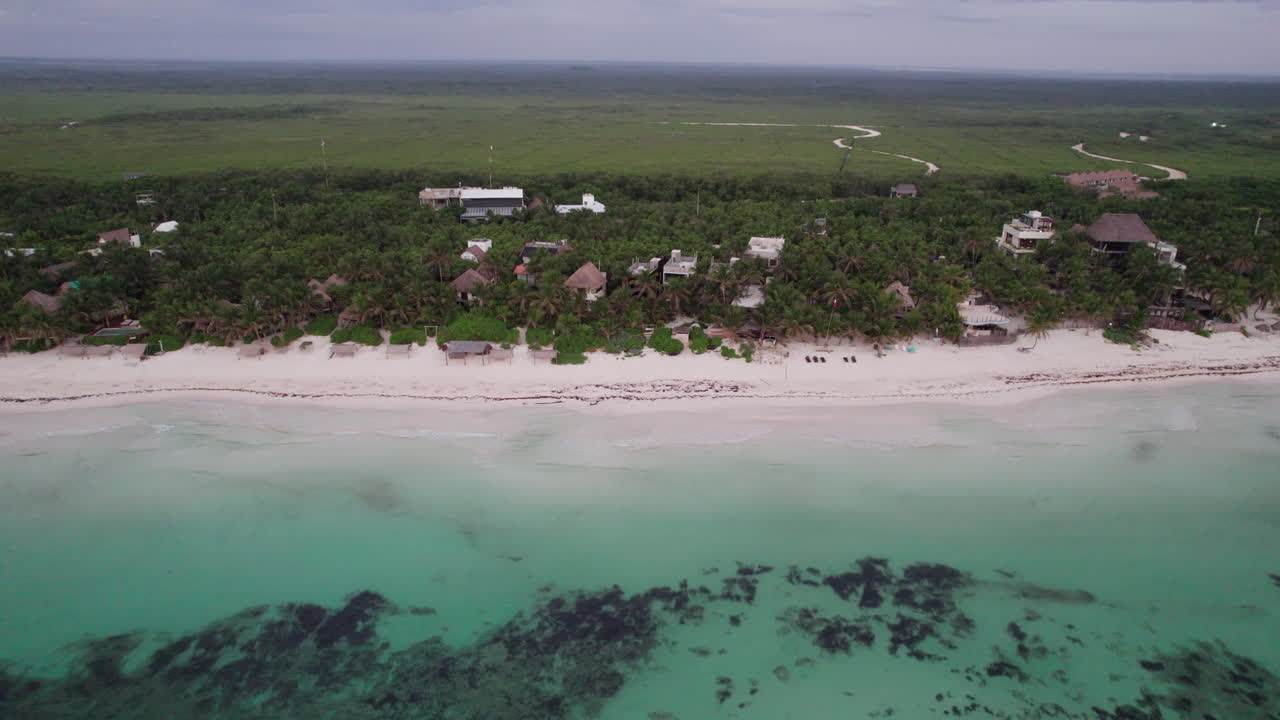 fotografía aérea de cabañas y chozas rodeadas de palmeras frente a una playa de arena blanca y un océano azul cristalino en tulum, méxico