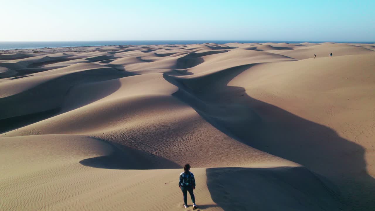 4K aerial drone flying over a female onlooker at Maspalomas sand dunes, Gran Canaria. Rolling golden dunes meet the ocean, highlighting the stunning beauty of the Canary Islands.
