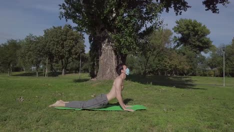 gran plano de órbita lenta de un joven europeo con máscara haciendo posturas básicas de yoga en la alfombra en la naturaleza