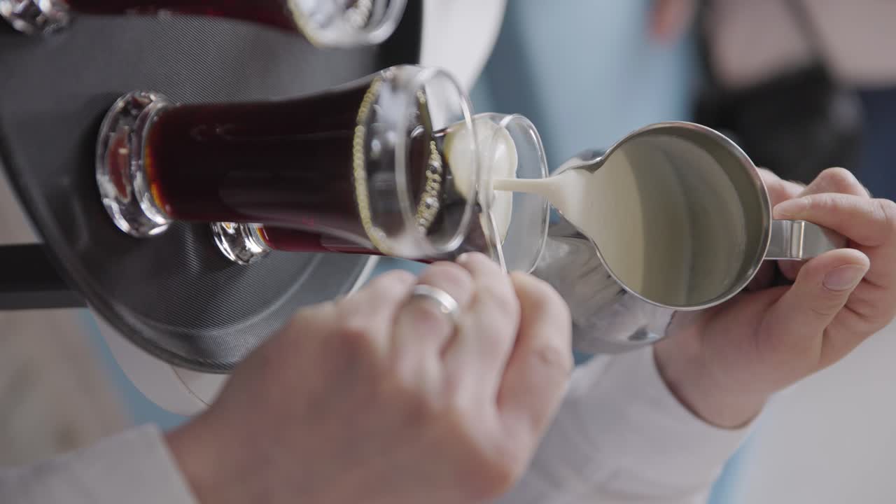 Barista uses spoon to taper flow and spread of cream or milk on dark coffee in glass