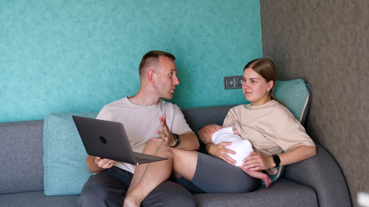 Caucasian family are in the room sitting on sofa. Parents of a newborn child looking at laptop in male arms.