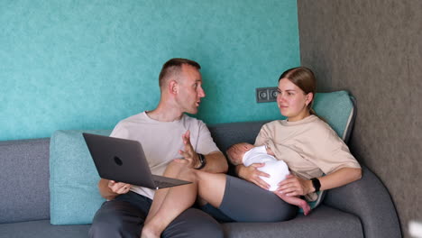 caucasian family are in the room sitting on sofa. parents of a newborn child looking at laptop in male arms.