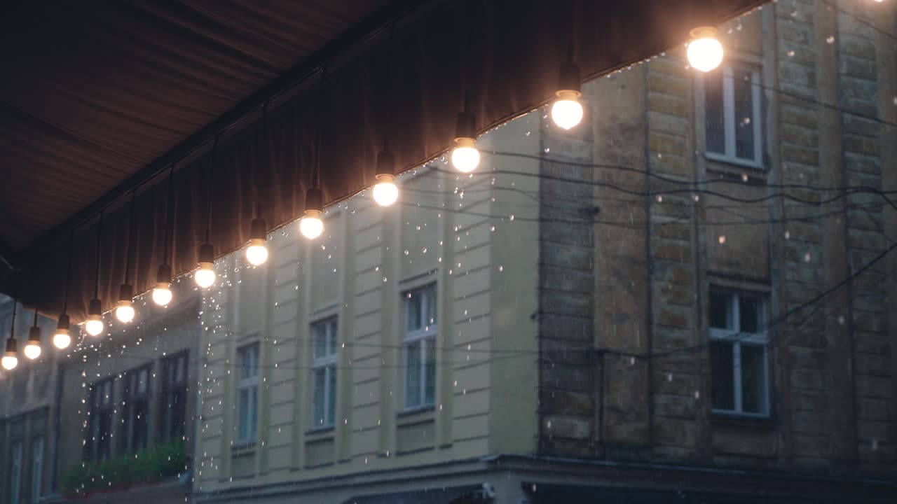 Regndroppar faller på restaurangens tak med en dekorativ rad lampor mot bakgrunden av en byggnad. Det regnar.