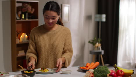 woman at home in kitchen preparing healthy vegetarian or vegan meal sprinkling herbs onto orzo pasta and roasted tomatoes